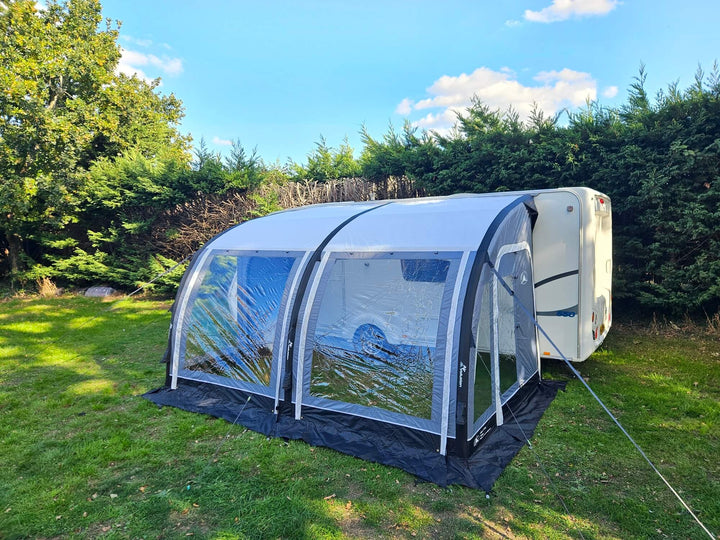 Rear angled view of the Sunncamp Ultima Versara Air 390 Deluxe Porch Awning attached to a caravan on a grassy campsite with trees and blue sky in the background.