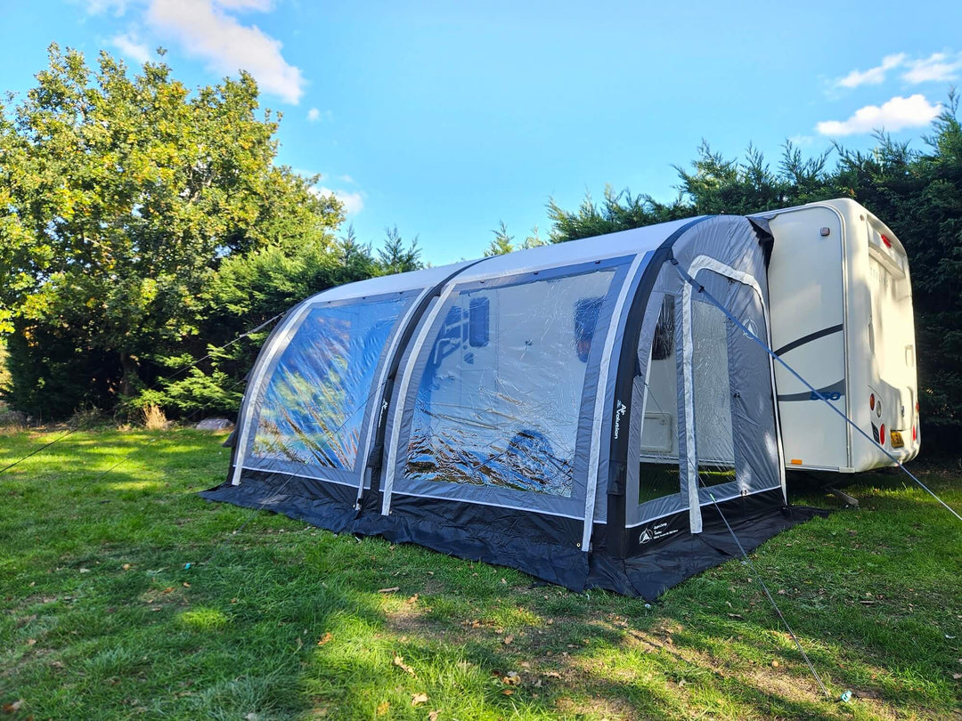 Angled side view of the Sunncamp Ultima Versara Air 390 Deluxe Porch Awning attached to a caravan on grass with blue sky and trees in the background.