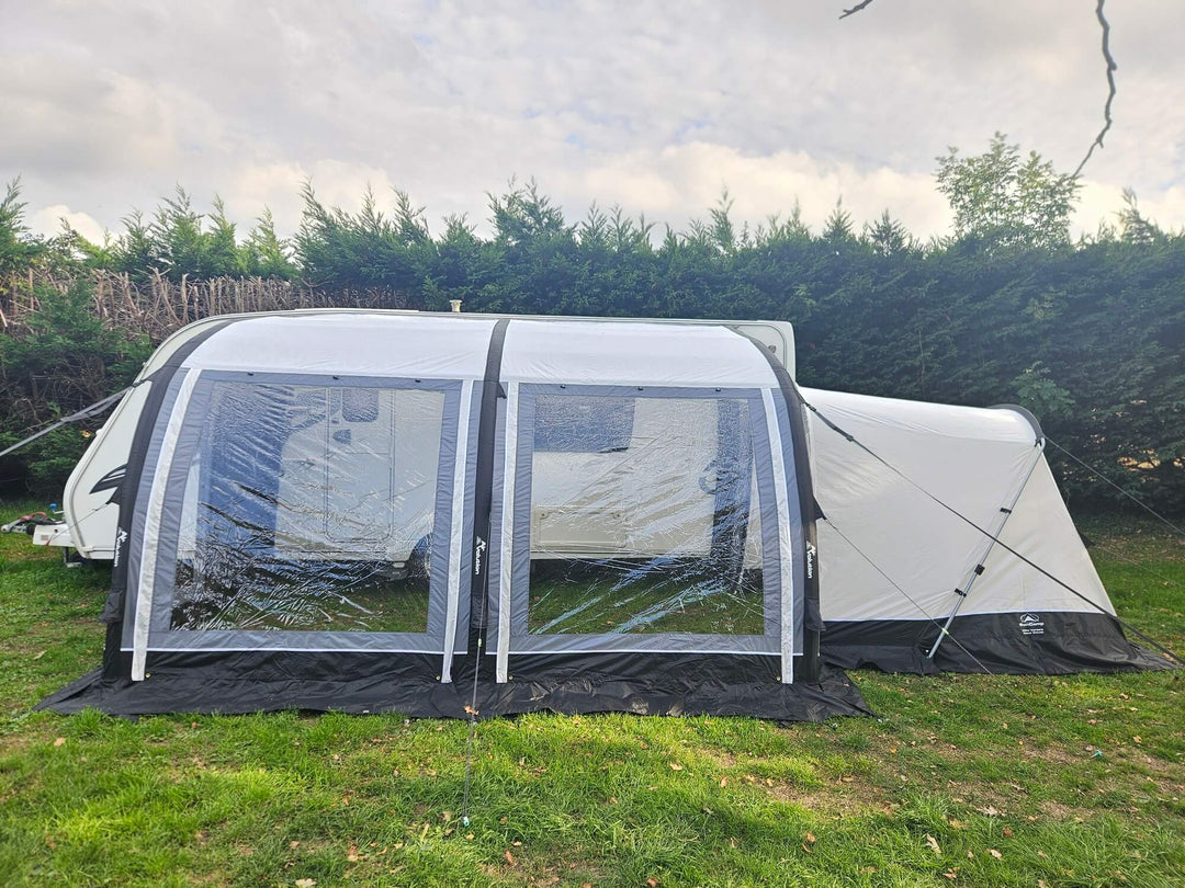 Front view of the Sunncamp Ultima Versara Air 390 Deluxe Porch Awning with large panoramic windows attached to a caravan on a grassy campsite with hedges and cloudy sky in the background.