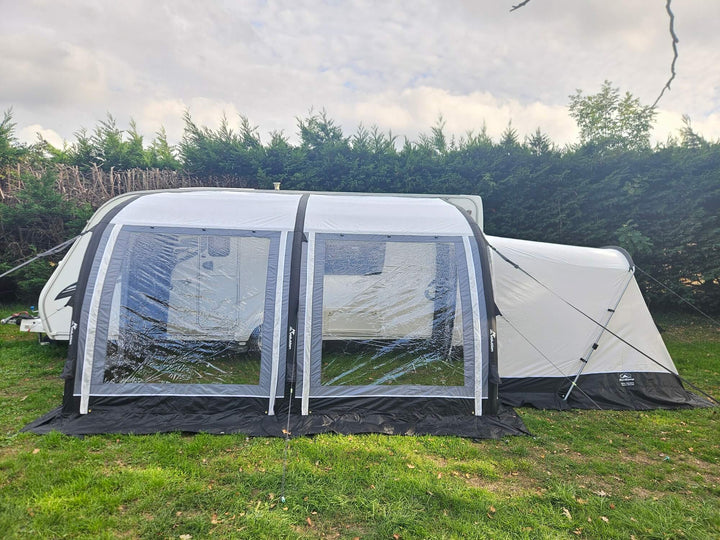 Front view of the Sunncamp Ultima Versara Air 390 Deluxe Porch Awning with large panoramic windows attached to a caravan on a grassy campsite with hedges and cloudy sky in the background.