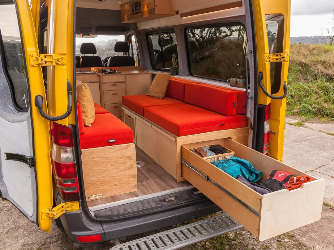 Campervan “Swale” Benches rear view with long pull-out storage drawer extended from red bench seat inside yellow campervan, countryside background.