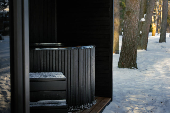 Taurus Modular Air Outdoor Sauna with black cold plunge tub and step stool on a wooden deck surrounded by snow and trees.