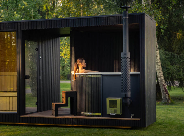 Taurus Classic Outdoor Sauna with integrated hot tub and chimney installed in a forest garden setting with trees and grass in the background.