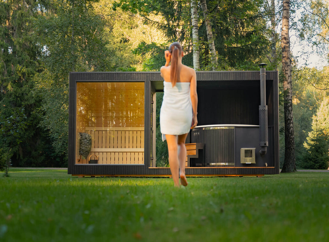 Taurus Grand Outdoor Sauna lifestyle image with a woman walking toward the unit on a grassy lawn surrounded by trees.