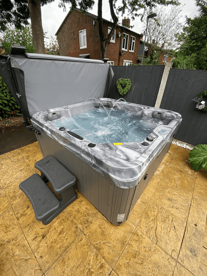 Luso Spas The Hawaii 2 Hot Tub bubbling on a yellow marble-patterned tiled patio with black fence and nearby trees in the background.