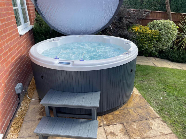 Outdoor setup of Luso Spas The Zodiac Plug & Play Hot Tub beside a brick wall with grey steps and bubbling water under an open cover.