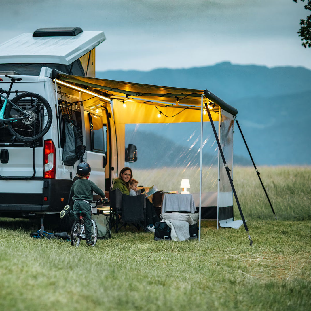Thule awning extended on a campervan with family camping in a grassy field at dusk.