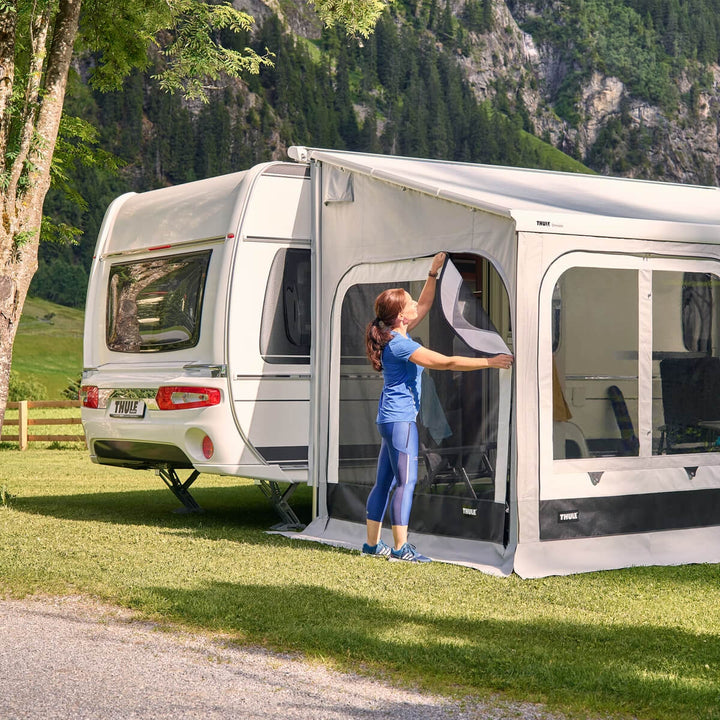 Woman opening an awning room door attached to a caravan on a grassy campsite with mountains.