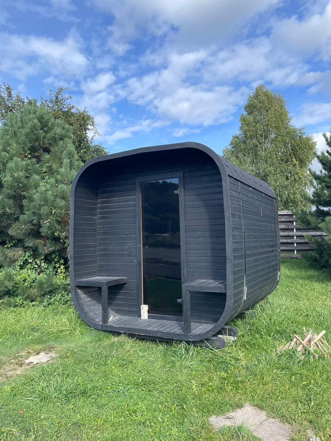 Ula Classic 6-Person Outdoor Sauna in black finish placed on grass in a garden setting with trees and blue sky background.