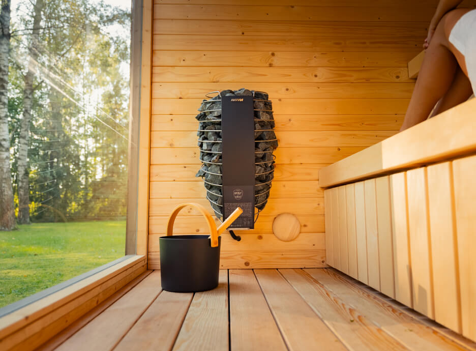 Interior of the Ula Classic Outdoor Sauna featuring a wall-mounted Harvia heater with sauna stones, wooden bench seating, and large glass window overlooking a garden.