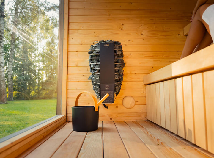 Interior of the Ula Classic Outdoor Sauna featuring a wall-mounted Harvia heater with sauna stones, wooden bench seating, and large glass window overlooking a garden.