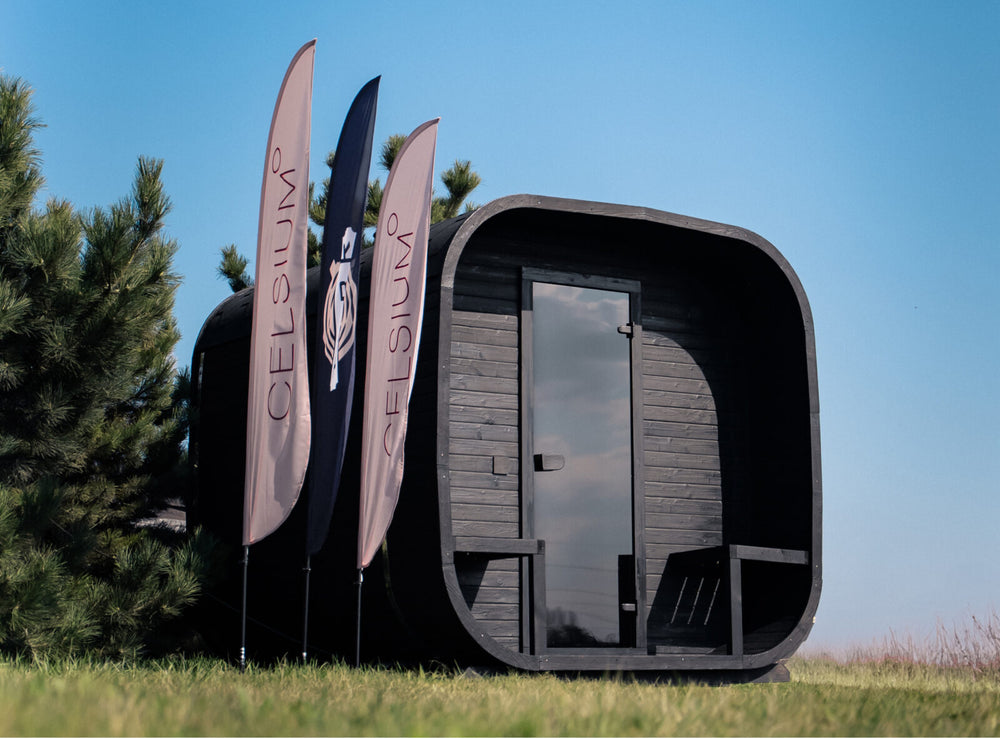 Ula Grand Outdoor Sauna in ebony finish placed on grass outdoors with trees and promotional flags in the background.