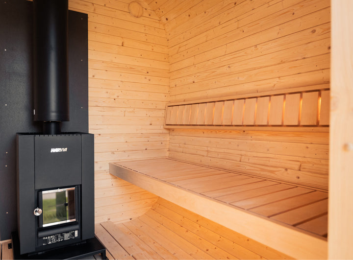Interior view of the Ula Grand Outdoor Sauna featuring wooden bench seating and a black Harvia heater inside a light timber-lined cabin.