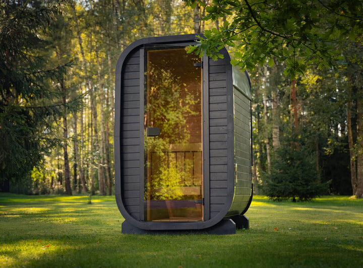 Ula Micro 2-Person Outdoor Sauna front view with curved black frame and glass door positioned on grass in a sunlit forest background.