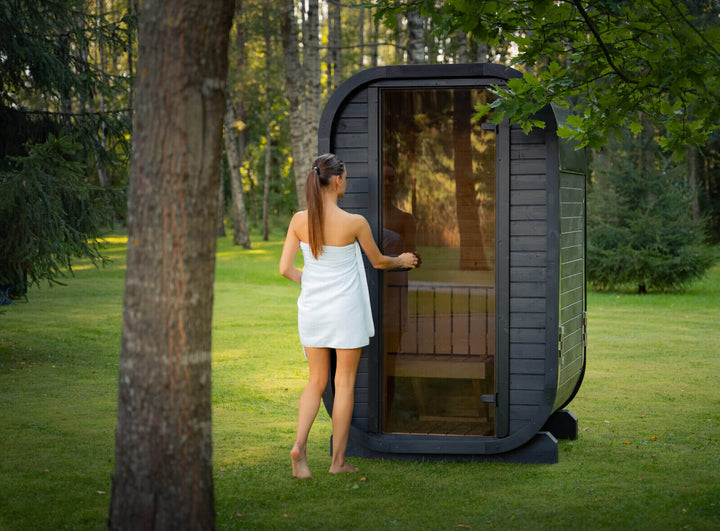 Ula Micro 2-Person Outdoor Sauna lifestyle view featuring a woman wrapped in a towel opening the glass door in a wooded garden setting.