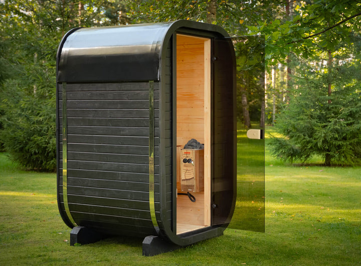 Ula Micro 2-Person Outdoor Sauna with open door showing wooden interior and heater inside, set on a grassy lawn with trees in the background.