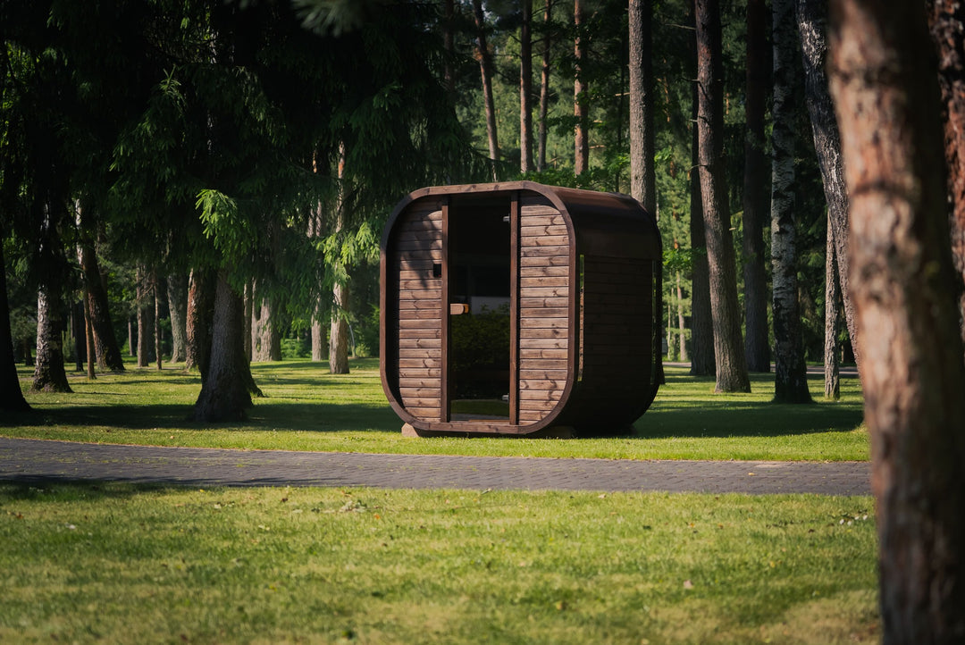 Ula Mini Outdoor Sauna exterior front angle view with curved wooden design and glass door in a park setting with trees and grass background.