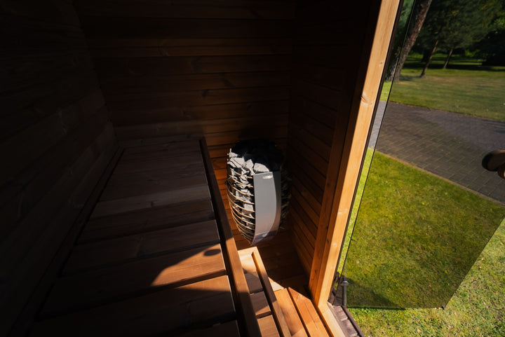 Ula Mini Outdoor Sauna exterior side view placed on grass in a wooded park setting with trees and pathway in the background.