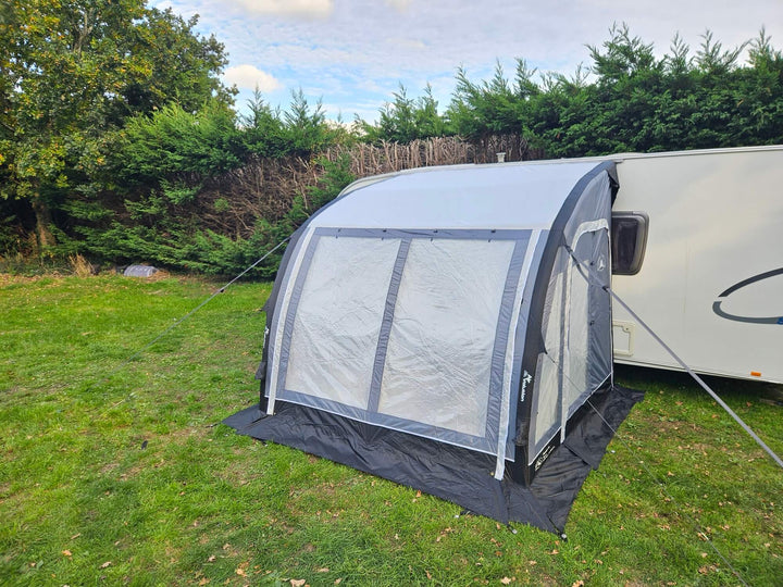 Front angled view of the Sunncamp Ultima Versara Air 260 Deluxe Porch Awning with large clear windows attached to a caravan on grass with trees and hedges in the background.