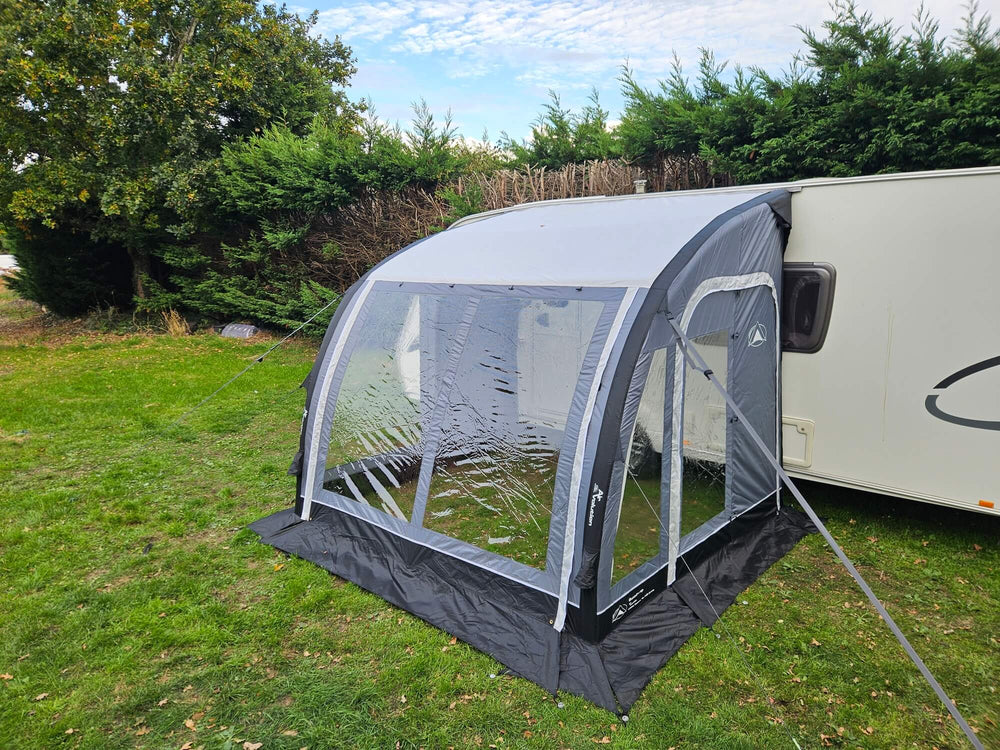 Angled side view of the Sunncamp Ultima Versara Air 260 Deluxe Porch Awning attached to a white caravan on a grassy campsite with hedge trees and cloudy sky in the background.