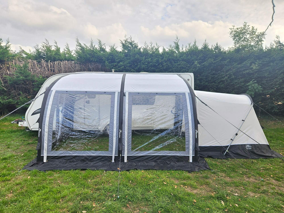 Front view of the Sunncamp Ultima Versara Air 390 Deluxe Porch Awning with clear window panels and rear extension attached to a caravan on grass with hedges in the background.