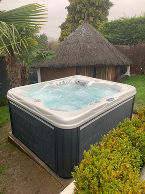 Angled outdoor view of Luso Spas The Vepo Hot Tub in operation with bubbling water and thatched hut in the background.