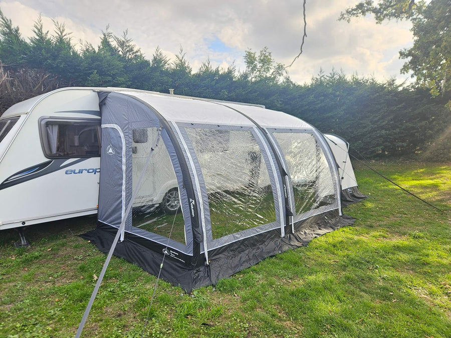 Sunncamp Ultima Versara Air 390 Deluxe Porch Awning attached to a white caravan on a grassy campsite with hedge trees and cloudy sky in the background.