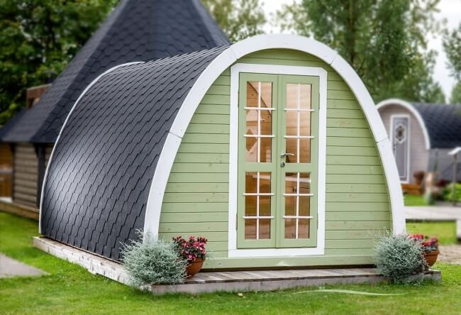 Viking Industrier 3.2 x 4.8m camping pod painted in sage green with white trim placed on a grassy yard with neighboring pods and cabins visible in the background.