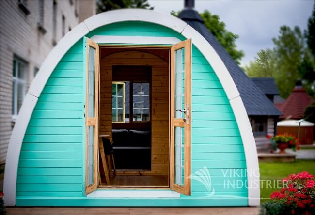 Viking Industrier 3.2 x 4.8m camping pod painted in teal with light wood frame shown in an outdoor setting with blurred buildings in the background.

