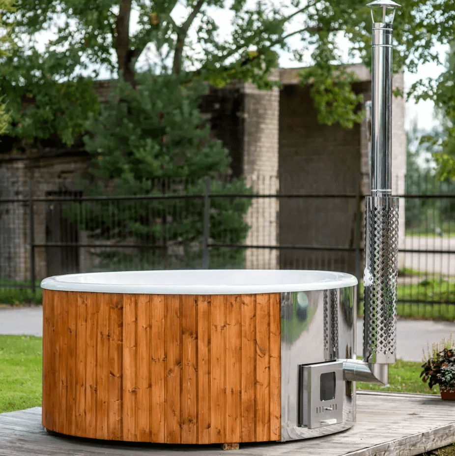 Viking Industrier Fiberglass Hot Tub with integrated stainless steel heater on a wooden platform, with a grassy lawn and metal fence in the background and large leafy trees behind.