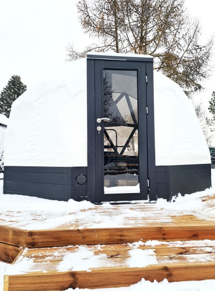 Front view of Viking Industrier Glamping Dome 10m with black door, snow-covered exterior, and wooden stairs, outdoors in winter.