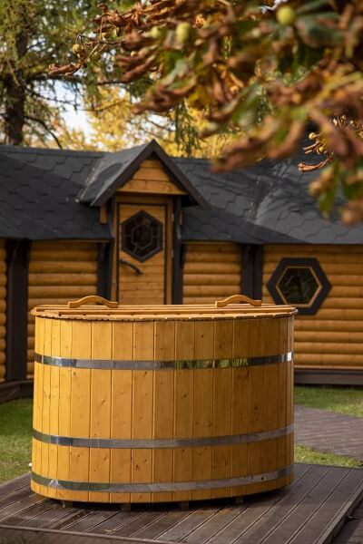Viking Industrier oval cold tub with closed lid on a wooden deck, placed in front of a wooden log cabin with decorative windows and trees in the background.