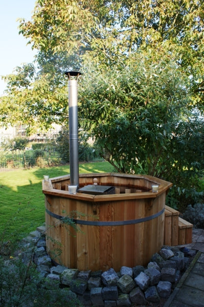 Viking Industrier Thermowood Hot Tub with a closed lid and a tall metal chimney, located on a stone base in a lush garden with trees.