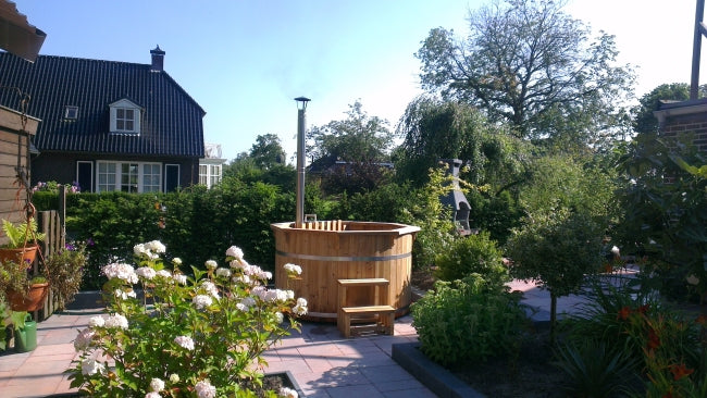 Viking Industrier Thermowood Hot Tub with a chimney in a sunny garden surrounded by flowers and shrubs, set on a tiled patio.