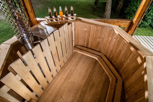 Interior view of a Viking Industrier Thermowood Hot Tub with wooden benches and metal stove, placed on a wooden deck with trees and grass in the background.