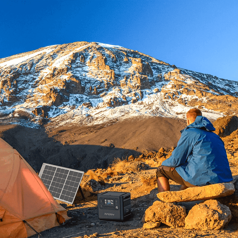 Aferiy P10 2400W Solar Generator Kit on rocky terrain with a camper sitting beside an orange tent, solar panel catching morning sunlight.