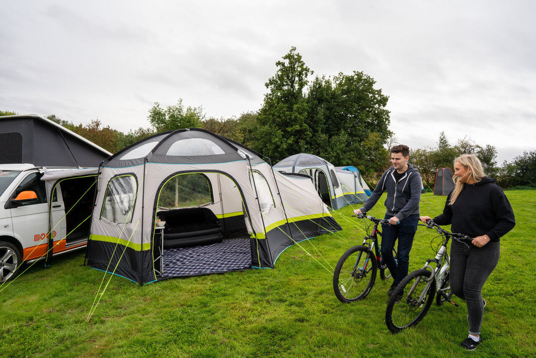 OLPRO Hive Campervan Awning pitched beside a campervan on a grassy campsite with two people riding bicycles in the foreground.