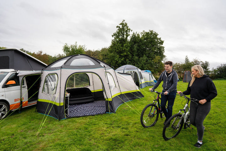 OLPRO Hive Campervan Awning pitched beside a campervan on a grassy campsite with two people riding bicycles in the foreground.
