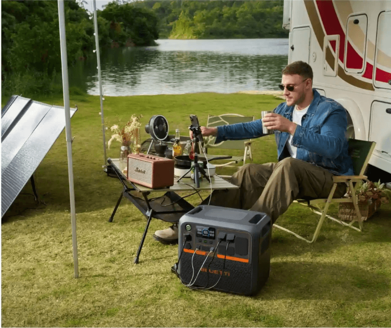 Man sitting by a camper van near a lake using the Bluetti AC200P power station connected to solar panels and electronic devices outdoors.