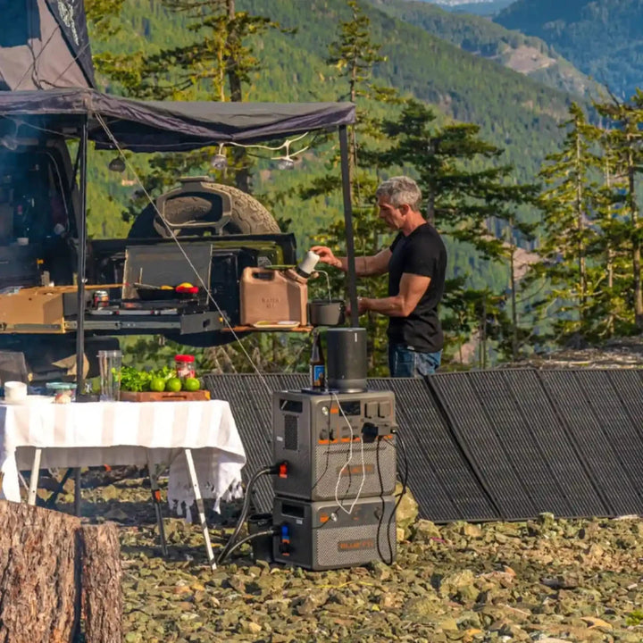 Man using a Bluetti AC240P power station outdoors at a camping site, surrounded by solar panels and cooking setup.
