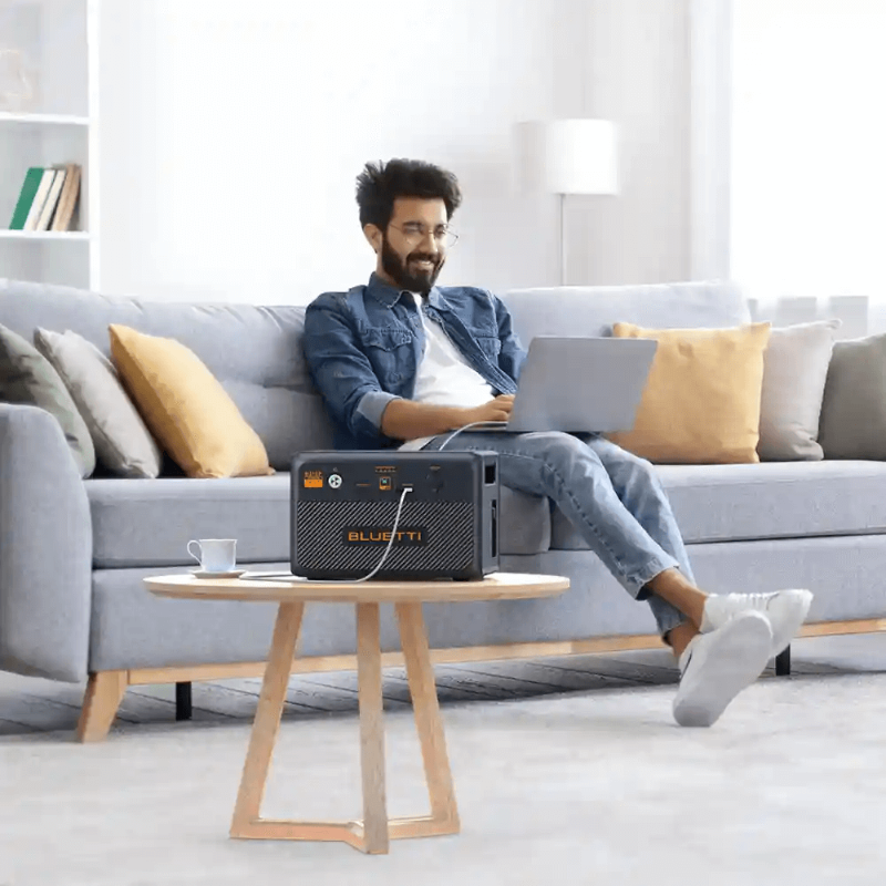 Man using a laptop in a living room with a Bluetti B210P battery unit powering devices.