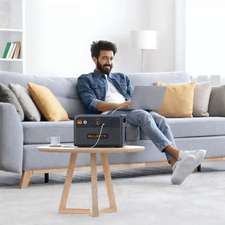 Man using a laptop in a living room with a Bluetti B210P battery unit powering devices.