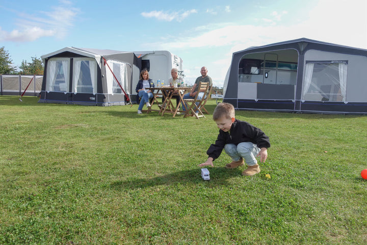 Family relaxing outside the Camptech Moto Windsor caravan awning on a grassy campsite under a blue sky.
