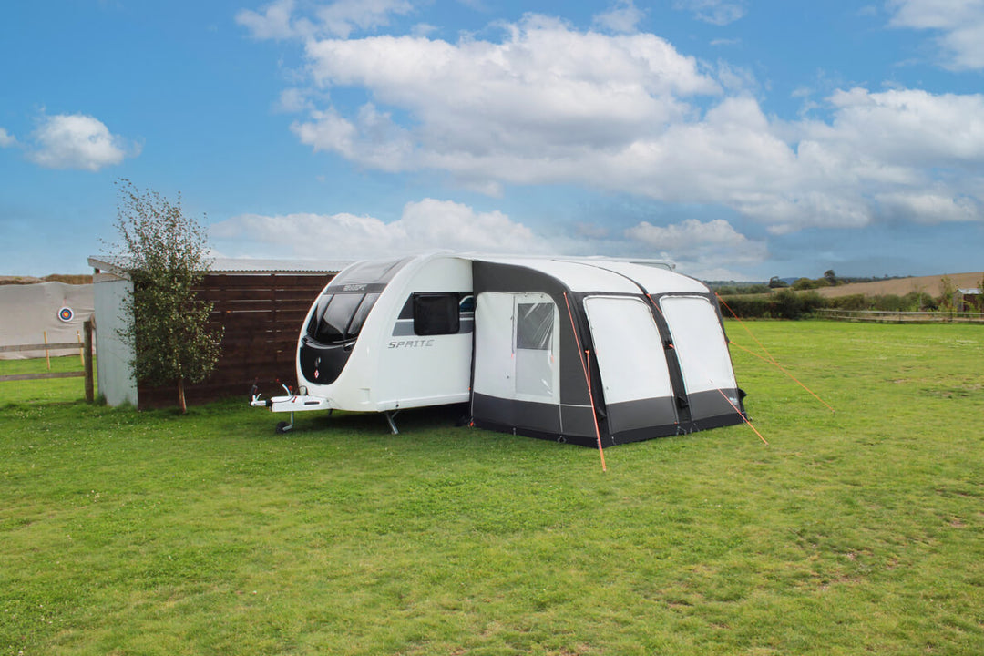 Camptech Starline 300 inflatable caravan awning beside a white Sprite caravan on an open grassy campsite with blue sky background.