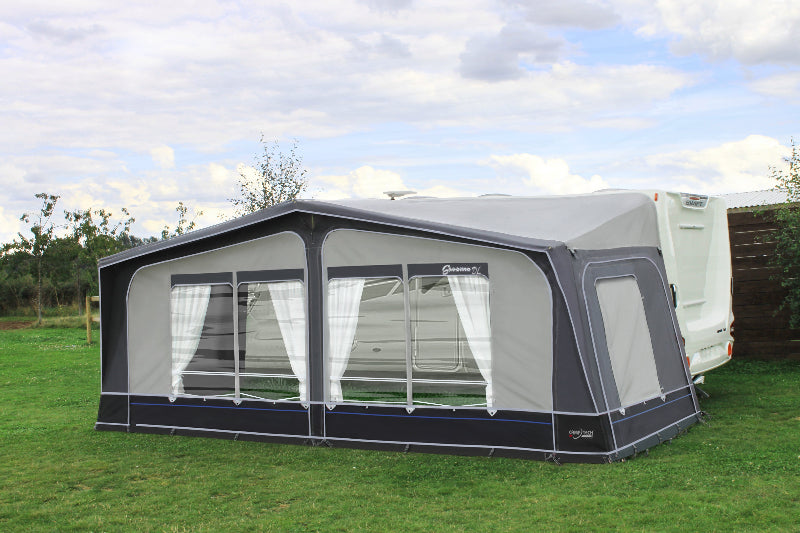Camptech Savanna DL caravan awning with large panoramic windows and white curtains, pitched on a grassy campsite beside a Swift caravan under a blue sky.