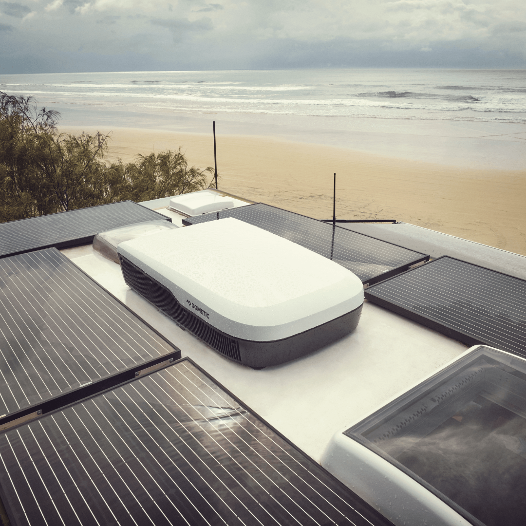 Top-down outdoor view of a Dometic FreshJet FJX7 3000 installed on an RV roof alongside solar panels. The beach and ocean are visible in the background under cloudy skies.