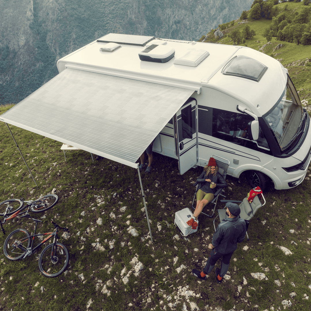 Overhead photo of a camper van parked on a grassy mountain ridge with the Dometic FreshJet unit installed on the roof; two people relaxing under the awning with mountain bikes nearby.