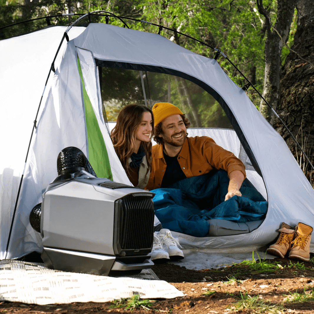 EcoFlow WAVE 3 air conditioner outside a camping tent, with a man and woman smiling from inside the tent, surrounded by greenery and camping gear.