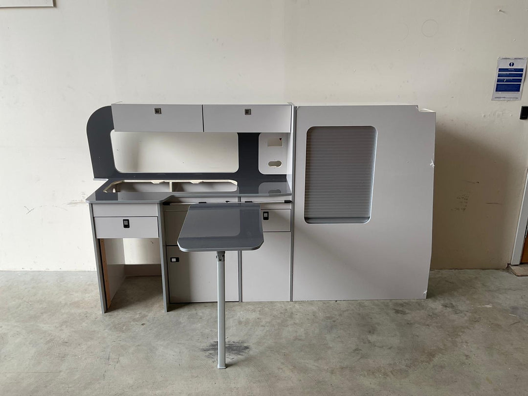 Ford Transit Custom Campervan Kitchen Unit Bespoke in dark grey finish with contrasting splashback and fold-out table, freestanding on artificial grass showroom flooring with display stands in the background.