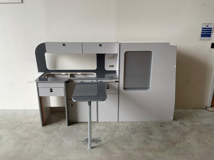 Ford Transit Custom Campervan Kitchen Unit Bespoke in dark grey finish with contrasting splashback and fold-out table, freestanding on artificial grass showroom flooring with display stands in the background.